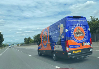 A blue and orange delivery van with business branding drives on a multi-lane highway under a partly cloudy sky.